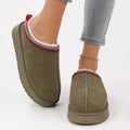 Women's faux suede slippers in olive with fleece lining and decorative woven trim worn with grey jeans
