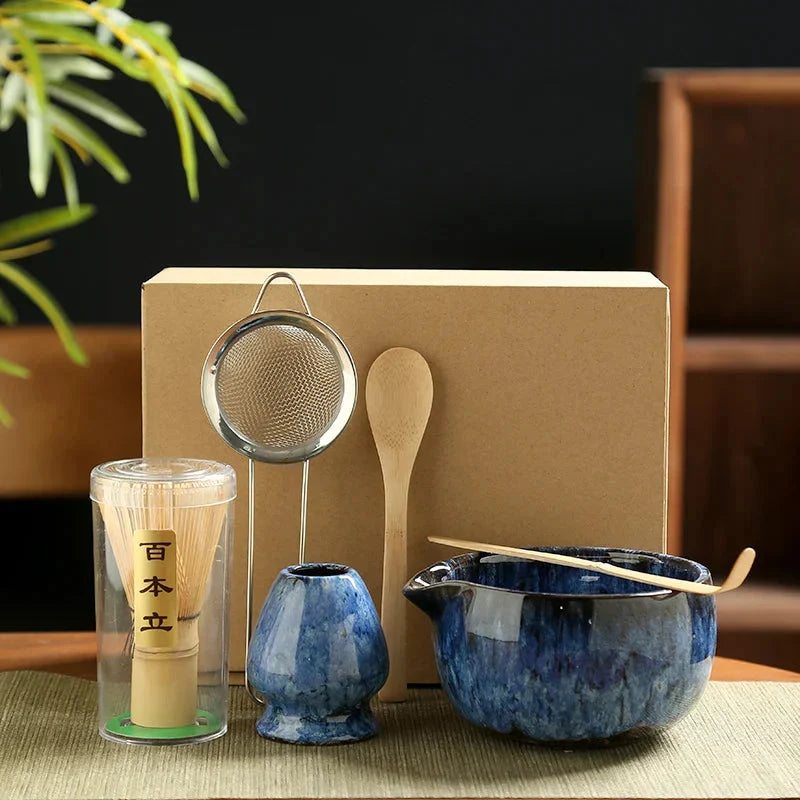 Six-piece matcha tea set with blue ceramic bowl, bamboo whisk, sieve, and spoon on woven mat