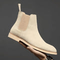 Men's Chelsea boots in beige with elasticated side panels held in hand against grey background