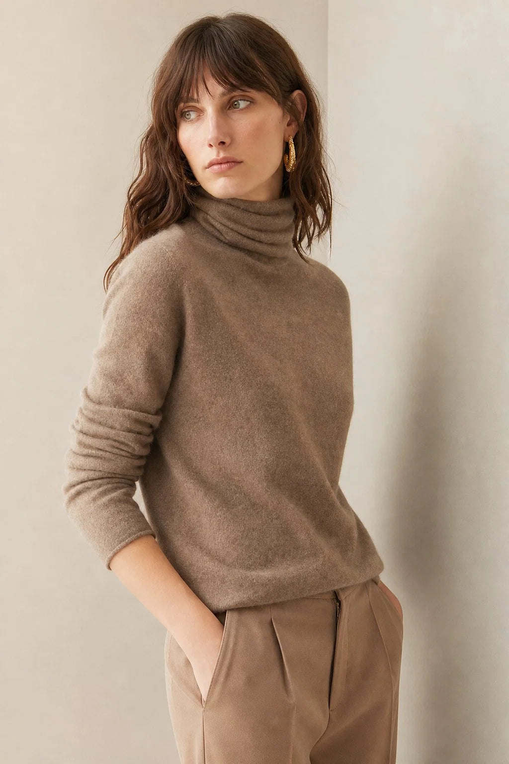 Woman wearing a taupe cashmere turtleneck jumper with long sleeves and a relaxed fit, styled with neutral trousers