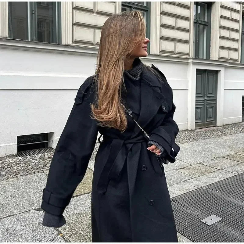 Long black trench coat with self-tie belt cinched at waist over denim jeans