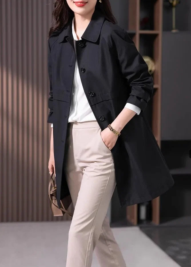 Women's short trench coat in black with notched collar and button closure worn over blouse and trousers